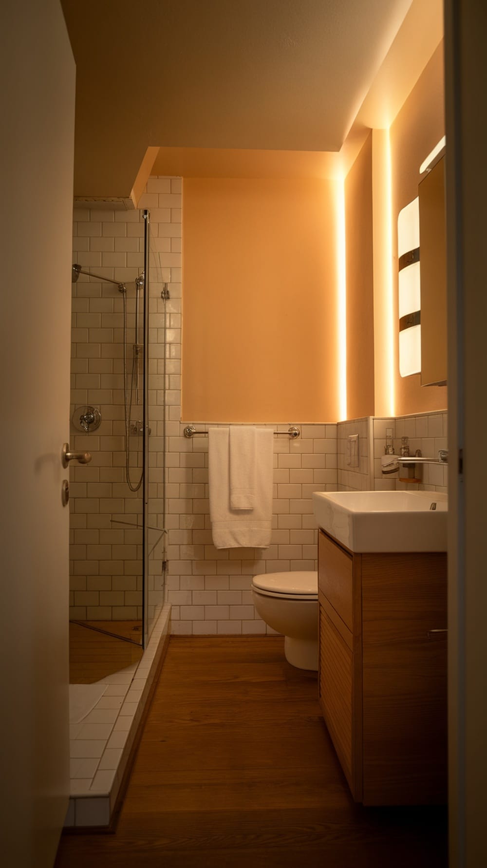 A small ensuite bathroom featuring soft, warm lighting, a glass shower, white tiles, and wooden accents.